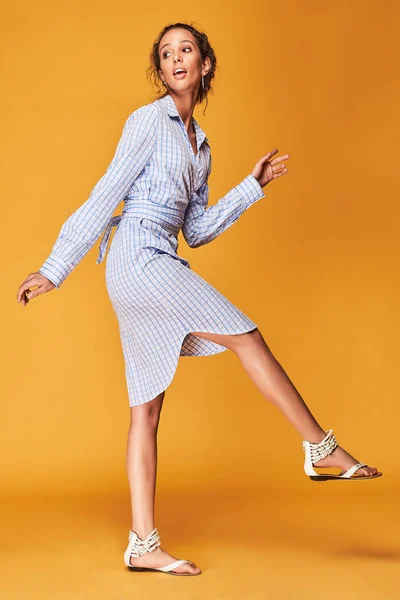 Full length portrait of tanned female model with curly brown hair, dressed in blue plaid shirt dress. Dynamic poses. Orange background