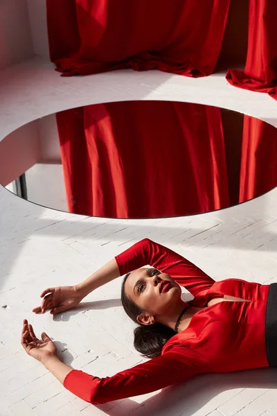 エレガントでセクシーな美しいブルネットの若い女性大きな丸い鏡の近くの床の上に敷設赤い絹のようなドレス 背景に赤のカーテン — ストック写真