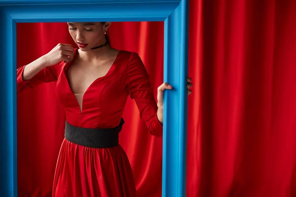ブルー フレームの後ろに立って長い赤いドレスの若いセクシーな女性 背景に赤のカーテン — ストック写真