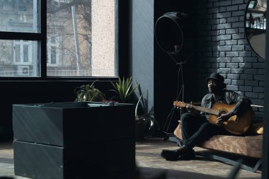 Müzisyen siyah palto ve yatağın üstüne oturan ve gitar çalmak kova şapka. Çatı odasında söz yazarı. Moda 2019 