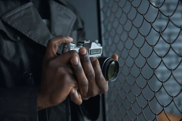 Vintage camera in hands. Man in black coat near chain link