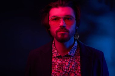 Neon ışık ile karanlık orta uzunlukta saç, bıyık ve sakal güneş gözlüğü, koyu renk ceketli ve çiçekli gömlek renksizdir yakışıklı erkek model portresi.