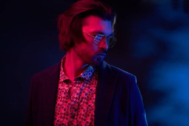 Neon ışık ile karanlık orta uzunlukta saç, bıyık ve sakal güneş gözlüğü, koyu renk ceketli ve çiçekli gömlek renksizdir yakışıklı erkek model portresi.