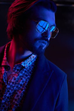 Neon ışık ile karanlık orta uzunlukta saç, bıyık ve sakal güneş gözlüğü, koyu renk ceketli ve çiçekli gömlek renksizdir yakışıklı erkek model portresi.