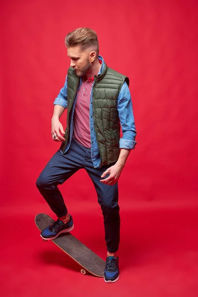 スケートボードに立つカジュアルなスタイルのファッショナブルな男 スタジオショット レッドバックグラウンド — ストック写真