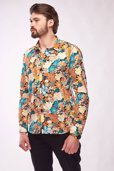 Fashion portrait of young male model with dark hair and beard, posing in floral shirt and black pants. White background