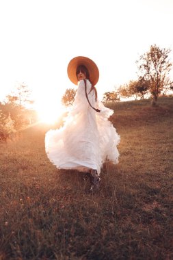 Beyaz elbiseli kadın ve gün batımında sahada yürüyen şık şapka. Altın saat. Moda.