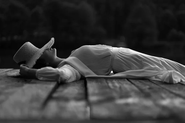 黑白梦幻的肖像 穿着复古衣服和草帽的女人 躺在池塘附近的码头上 — 图库照片