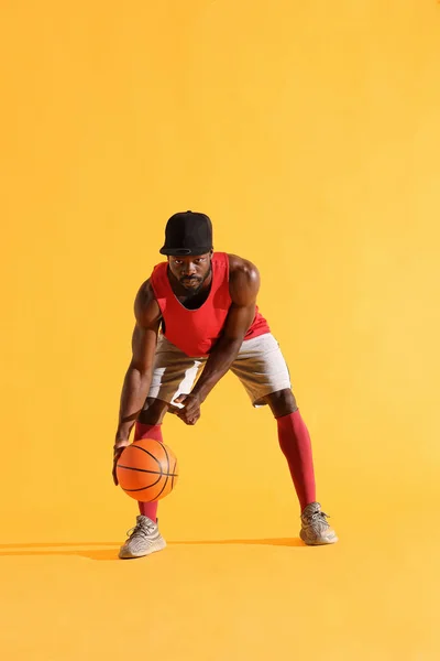 Full length portrait of black man in red shirt, black cap and grey shorts playing basketball. Studio, yellow background