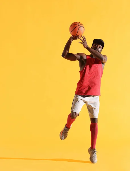 Full length portrait of black man in red shirt, black cap and grey shorts playing basketball. Studio, yellow background