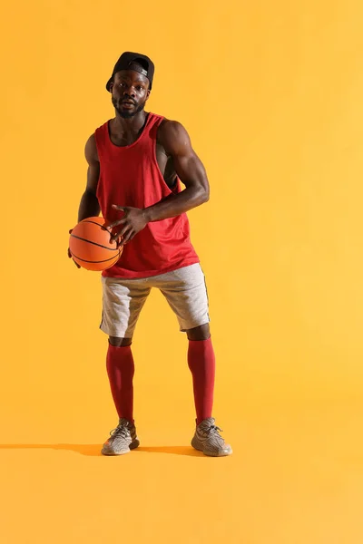 Full length portrait of black man in red shirt, black cap and grey shorts playing basketball. Studio, yellow background