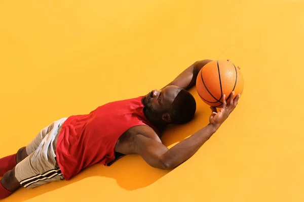 Portrait of black man in red shirt and grey shorts laying on the floor with basketball ball. Studio, yellow background