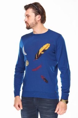 sakallı ve koyu saçlı bir adam beyaz bir arka plan üzerinde tüyleri ve koyu kot ile mavi bir kazak üzerinde duruyor elinde altın bir saat var ve sol görünüyor