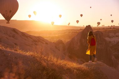 Kapadokya 'da gökyüzünde hava balonları ve kırmızı çantalı bir kız ile gündoğumu fotoğrafı. Koyu renk kot, sarı hırka ve beyaz spor ayakkabılar tepede durup manzarayı izliyor.