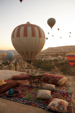 Büyük kırmızı ve beyaz balon mavi gökyüzünde, kumlu bir tepenin üzerinde bir balon daha yükseliyor. Yastıklı bir Türk halısı, bir tabak meyve ve iki balkabağı ve halının ortasında bir masa var.