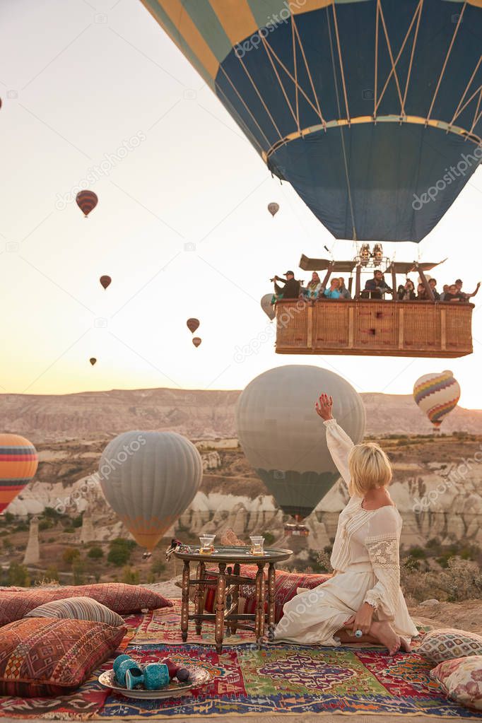 a girl in white dress sitting on old turkish carpet with pillows and table with tea waving to people in a big air balloon in the sky in Cappadocia