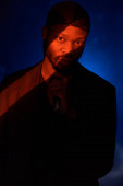 pohledný muž tmavé pleti v šátku, černé klasické bundě a tričku s mikrofonem v ruce, stojí ve stínu na modrém pozadí a dívá se do kamery