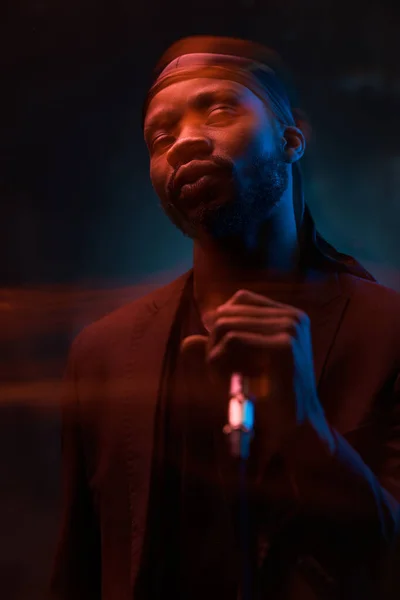 dark-skinned handsome guy in a bandana, black classic jacket and t-shirt holds a microphone in his hands and emotionally sings in a dark studio in red light