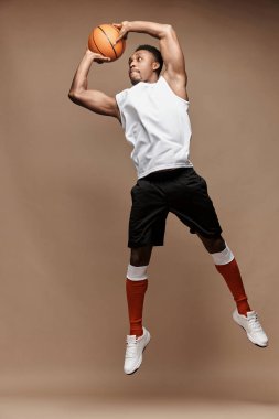 Stüdyoda koyu tenli bir atletizm oyuncusunun beyaz tişört, siyah şort, kırmızı uzun çorap ve beyaz spor ayakkabı giyerek bej bir arka plan atlamasıyla tam boy fotoğrafı.