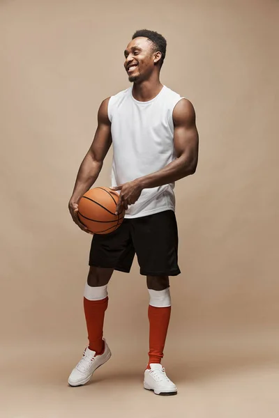 full length photo of a black athletic basketball player in the studio on a beige background wearing a white T-shirt, black shorts, red long socks and white sneakers, he holds the ball and smiles