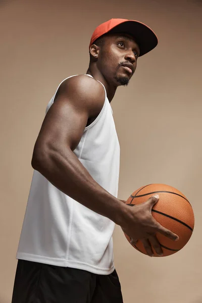 photo of a dark-skinned athletic basketball player in studio on a beige background posing with a ball, wearing a white t-shirt, black shorts, red long socks and a cap, he is looking up