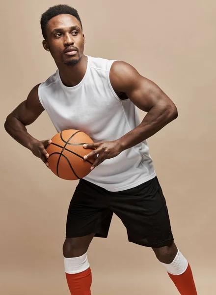 dark-skinned athletic basketball player in studio on a beige background posing with a ball wearing a white T-shirt, black shorts, red long socks