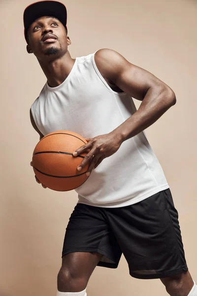 photo of a dark-skinned athletic basketball player in studio on a beige background posing with a ball, wearing a white t-shirt, black shorts, red cap and he is looking up