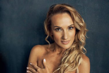 Uzun sarı kıvırcık saçlı, profesyonel makyajlı ve güzel kahverengi gözlü güzel bir kadının portresi, poz verir ve göğüslerini gizleyen kameraya bakar.