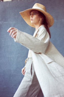 Gri bir arka planda gri saçlı, çıplak makyajlı, gri keten pantolon giymiş, kafasında saman şapkayla bej bluz ve pelerin giymiş güzel bir kızın portresi.