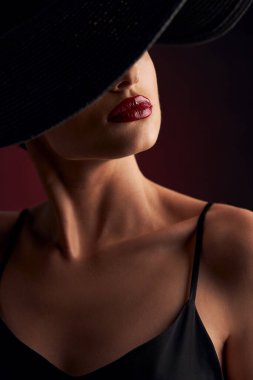 Profesyonel makyajla bronzlaşmış güzel bir kızın portresi, kırmızı dudaklar, koyu renk bir arka plan, siyah bir elbise ve gözlerini kapatan siyah bir şapka.
