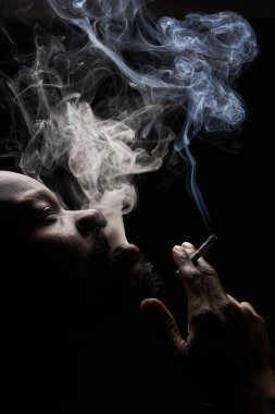 Koyu tenli, kaslı yakışıklı, siyah arka planda sakallı bir adamın profil fotoğrafı. Ağzından sigara solur ve yukarı bakar.