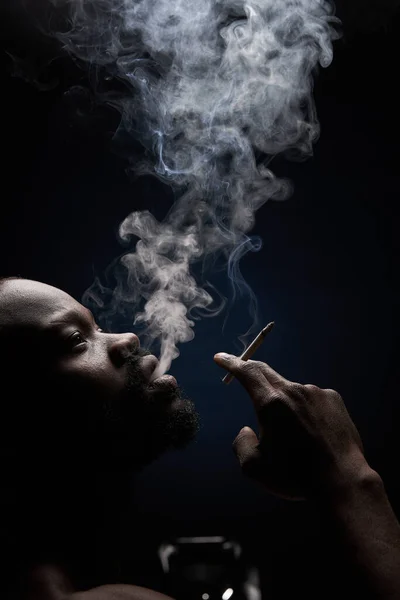 照片上是一个深色皮肤 体形英俊 留着胡子 背景深色的男人 手里拿着香烟 嘴里喷出浓烟 抬起头来看 — 图库照片