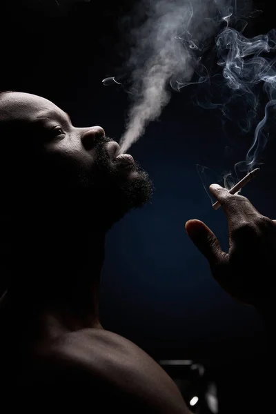 暗い肌をしたハンサムな男のプロフィール写真暗い背景にひげを生やした男彼は手にタバコで彼の口から煙を呼吸し 検索します — ストック写真
