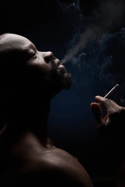 profile photo of a dark-skinned muscular handsome guy with a beard on a dark background, who breathes out smoke from his mouth with a cigarette in his hand and looks up