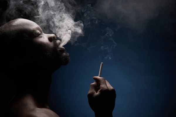 profile photo of a dark-skinned muscular handsome guy with a beard on a dark background who breathes out smoke from his mouth with a cigarette in his hand