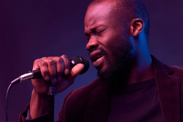 portrait of a dark-skinned handsome guy in dark jacket and t-shirt holds a microphone in his hands and emotionally sings in a dark studio with red and blue light
