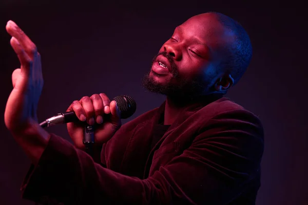 portrait of a dark-skinned handsome guy in dark jacket and t-shirt holds a microphone in his hands and emotionally sings in a dark studio with red and blue light