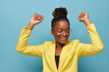 Sarı ceketli, şeffaf gözlüklü, koyu tenli güzel bir kızın portre fotoğrafı ellerini hareket ettiriyor, gülümsüyor ve aşağıya bakıyor.