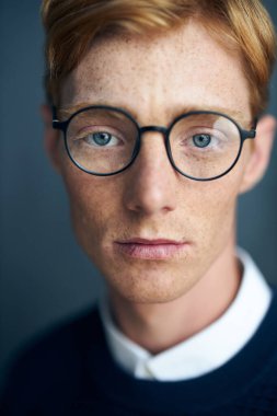 Koyu arkaplanda çilli, genç, kızıl saçlı bir adamın portresi. Beyaz bir gömlek üzerine koyu mavi bir süveter giyiyor ve siyah çerçeveli şeffaf gözlükleri var.