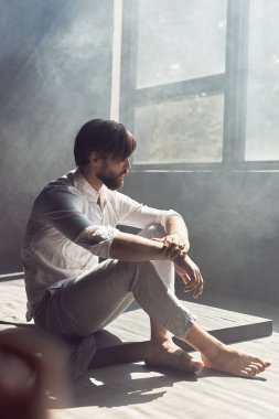 Kahverengi saçlı yakışıklı sakallı bir adamın fotoğrafı. Stüdyoda yerde sigara ve güneş ışığıyla oturuyor. Beyaz keten bir gömlek ve pantolon giyiyor ve başka tarafa bakıyor.