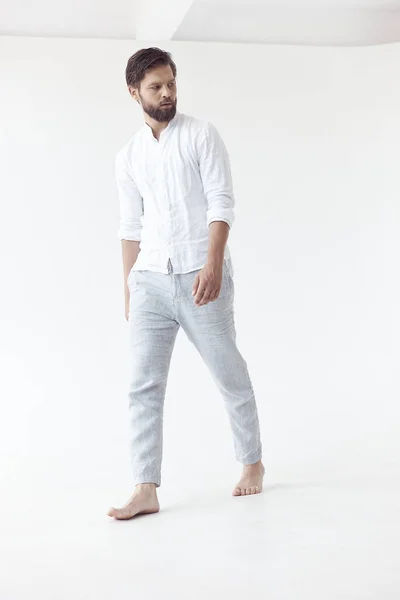 full-length photo in high key on a white background of a handsome bearded man with brown hair, he is wearing a white linen shirt and blue pants, bared feets, he looks away