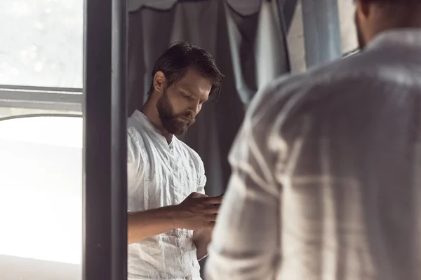 photo in the mirror of a handsome bearded man with brown hair, who stands and looks intently at the phone in his hands, he is wearing a white linen shirt