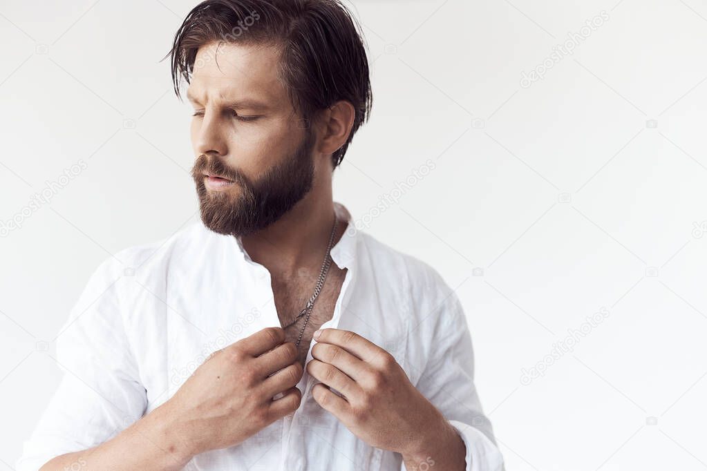 portrait photo of a handsome bearded man with brown hair on white background, he is wearing a white linen shirt, looks away and touches his shirt