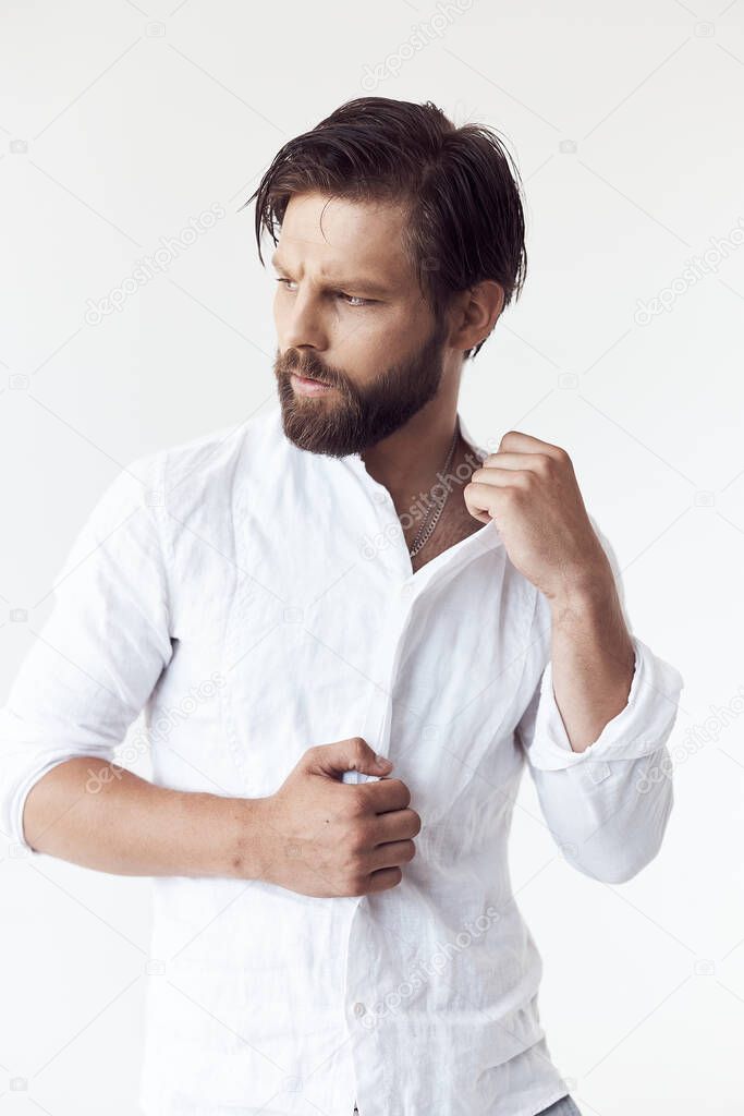 portrait photo of a handsome bearded man with brown hair on white background, he is wearing a white linen shirt, looks away and touches his shirt