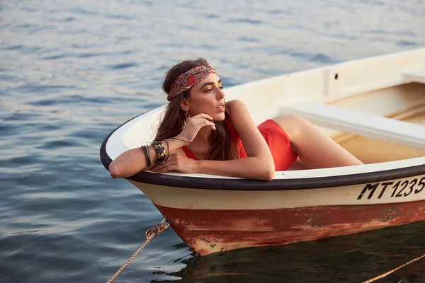 beautiful girl in a red swimsuit, headband, bracelets on her hands, she has a professional make-up and hair styling lying in a boat by the sea and posing