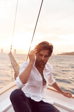 Yatta gün batımında sakallı yakışıklı bir adam, beyaz keten gömlek ve pantolon giymiş, vahşi bir ifadeyle saçlarını rüzgardan ayırıyor.