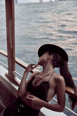 Güzel ve mutlu siyah saçlı kız, siyah bluzlu, pantolonu ve şapkası denizde giden bir geminin gövdesinde.