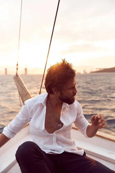 handsome man with a beard at sunset on a yacht wearing a white linen shirt and pants with a brutal expression
