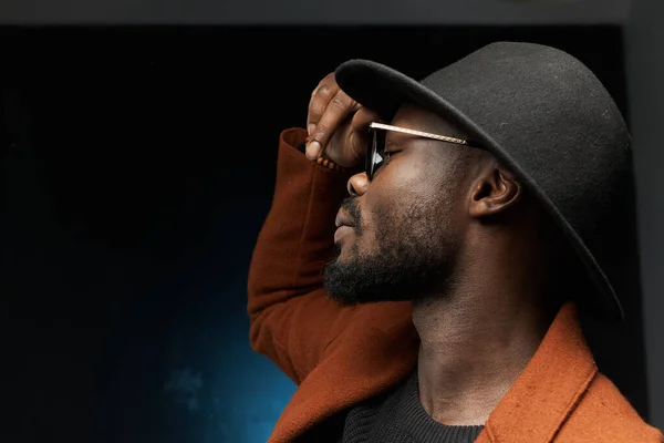 profile photo of a black man in a black hat, brown jacket and sweater on a dark background in the studio, he holds his glasses with one hand