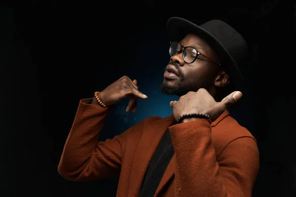 portrait photo of a black man with glasses, a black hat, a brown jacket and a jacket on a dark background in the studio, who faces the studio light and moves his hands